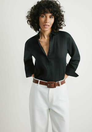 Kobieta o kręconych włosach nosi czarną koszulę zapinaną na guziki, białe spodnie i brązowy pasek, stoi na jednolitym jasnym tle.