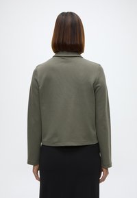 Χακί ριγωτό μπουφάν με γιακά, μακριά μανίκια και ε�υθεία βάση, φορεμένο πάνω από μια μαύρη φούστα. Ομαλή υφή, μινιμαλιστικός σχεδιασμός.