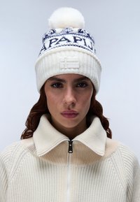 Berretto lavorato a maglia bianco con accenti blu, con un pom-pom sulla parte superiore, schemi sovrapposti e una finitura testurizzata. La parte anteriore mostra un patch con il logo.
