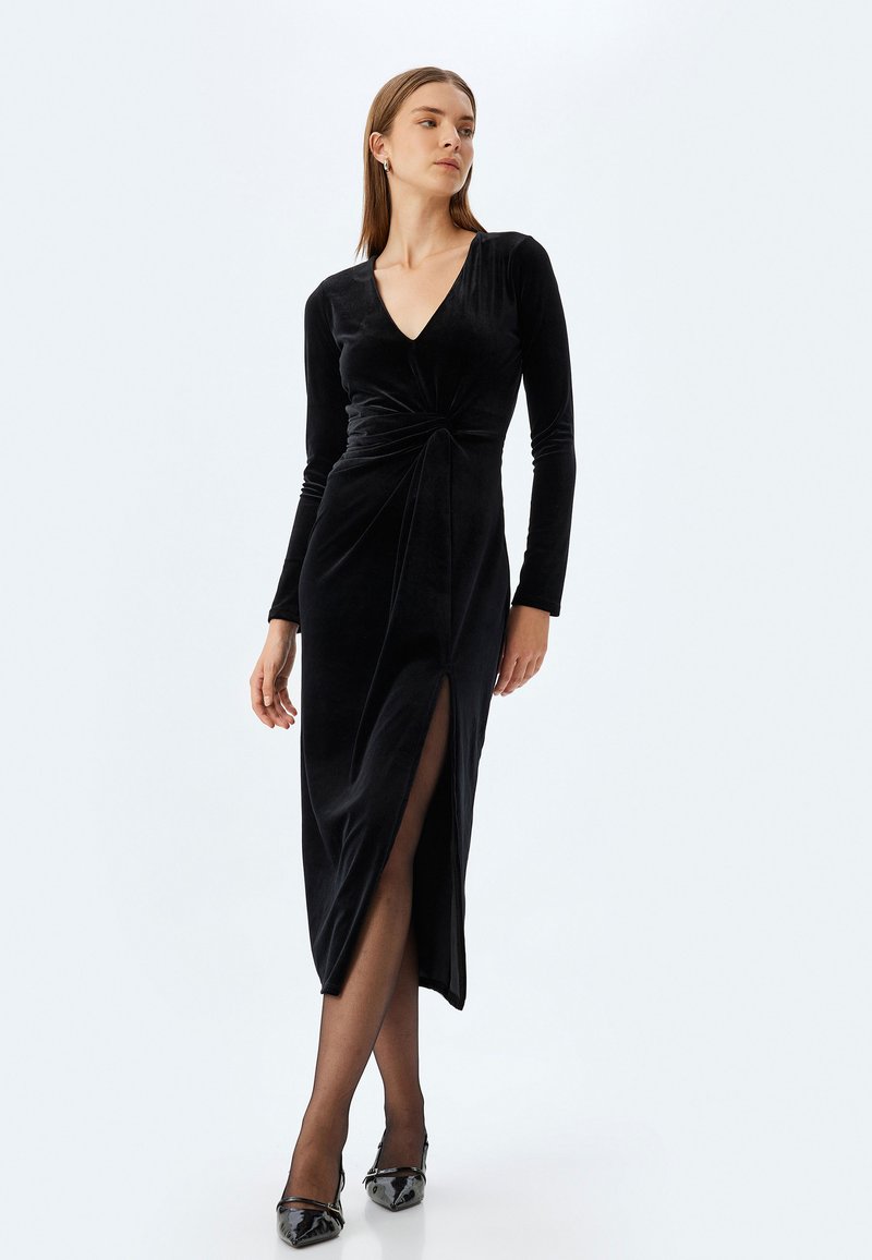Zwarte fluwelen jurk met lange mouwen en een V-hals, met een twistdetail bij de taille en een zijsplit voor gemakkelijke beweging.