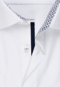 Camicia bianca con colletto a motivi blu navy, caratterizzata da un'apertura con un solo bottone e una texture sottile. Gli accenti includono cuciture blu navy.