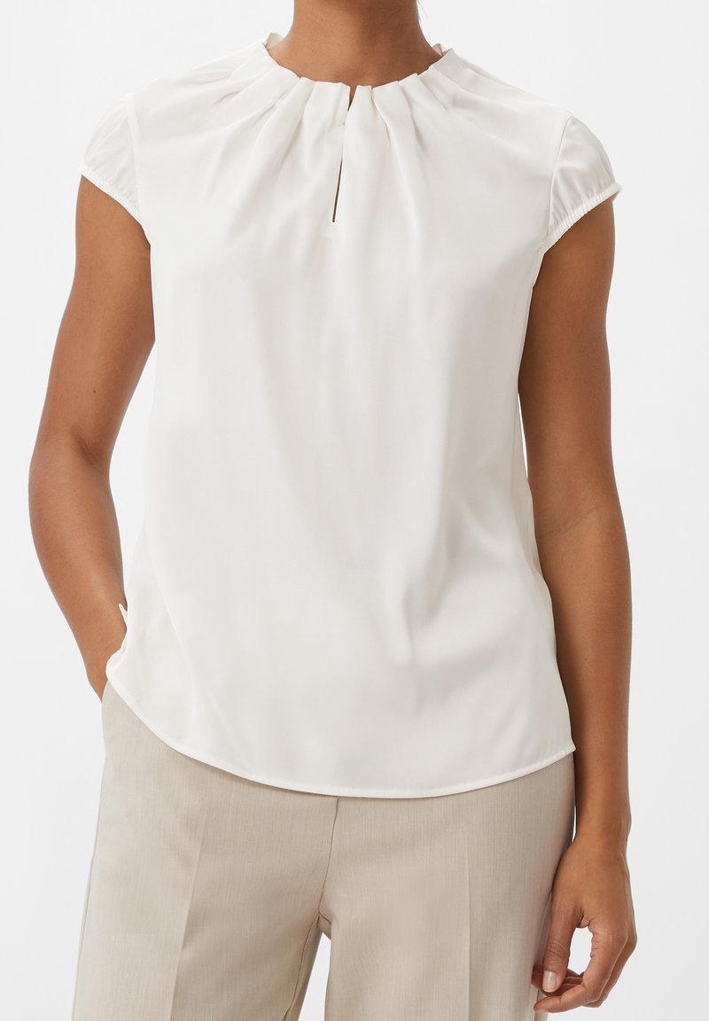 Femme portant un chemisier blanc sans manches avec un col froncé et un pantalon beige, mains détendues le long du corps sur un fond uni.