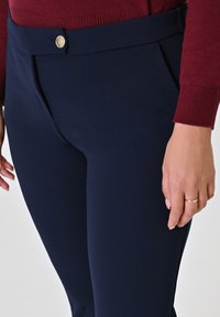 Pantaloni tailorizzati blu navy con una texture liscia, caratterizzati da un bottone dorato e una parte anteriore piatta. Tasche laterali visibili.