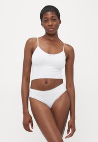Žena s kratkom kosom koja nosi bijeli teksturirani crop top i bijelo donje rublje s čipkastim rubom, stoji nasuprot jednostavne bijele pozadine.