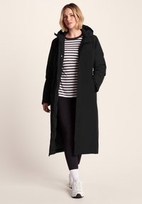 Woman walking wearing a long black hooded coat, black leggings, white sneakers, and a white and black striped shirt against a plain background.
