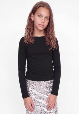 Une jeune femme aux longs cheveux bruns et ondulés pose sur un fond blanc. Elle porte un haut noir à manches longues et une jupe argentée à sequins.