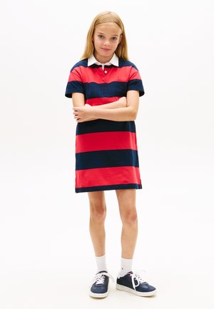 Młoda dziewczynka stoi z założonymi skrzyżowanymi rękami, ubrana w czerwoną i granatowo paskowaną sukienkę polo, białe skarpetki i granatowe trampki.