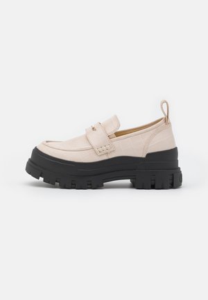 Chaussures synthétiques beiges à texture crocodile, dotées d'une semelle épaisse en caoutchouc noir et d'un design sans lacets avec une languette arrière pour faciliter l'enfilage.