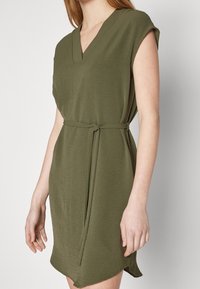 Olivově zelené šaty s krátkými rukávy, výstřihem do V a vázaným pasem. Měkká tkanina s hladkou texturou, s mírně zakřiveným lemem.