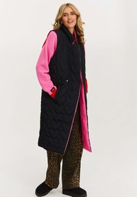 Czarny długi, pikowany kamizelka z różowym podszewką, noszona na różowym swetrze i spodniami w leopardzi wzór. Posiada zapięcia na guziki i kieszenie boczne.