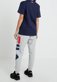 Osoba w granatowej koszulce i jasnoszarych dresach z czerwono-niebieskimi geometrycznymi wzorami, w czarnych sneakersach FILA, stojąca na tle białym.