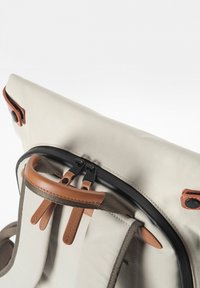 Close-up of beige backpack top with leather handle and zipper pulls, featuring black zipper and leather snap button details.