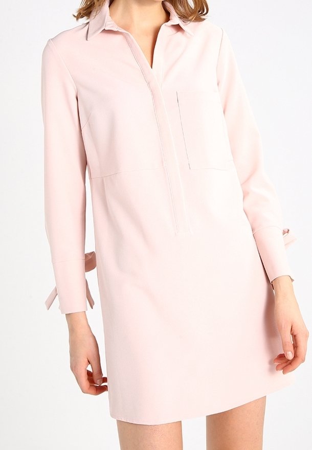 Femme portant une robe chemise manches longues de couleur rose pâle avec un col et un détail de poche avant, debout devant un fond blanc.