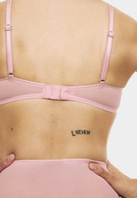 Lingerie rosa con un pannello posteriore trasparente, spalline regolabili e chiusura con ganci. Presenta bordi ondulati e un tatuaggio con la scritta "LUCIEN."