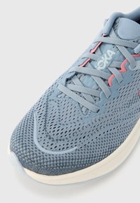 Αθλητικό παπούτσι σε ανοιχτό μπλε χρώμα με επάνω μέρος από δίχτυ, κείμενες λεπτομέρειες, ροζ πινελιές και επίπεδα κορδόνια. Στρογγυλεμένη λευκή σόλα για απορρόφηση κραδασμών και υποστήριξη.