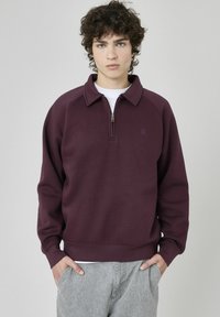 Sweatshirt bordeaux à col rond avec demi-zip, manches et ourlet côtelés. Tissu texturé avec un petit logo sur la poitrine. Pantalons gris visibles.