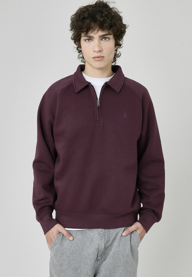 Sweatshirt bordeaux à col rond avec demi-zip, manches et ourlet côtelés. Tissu texturé avec un petit logo sur la poitrine. Pantalons gris visibles.