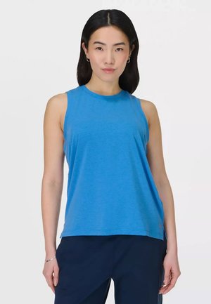 Vrouw met lang donker haar, gekleed in een mouwloze blauwe top en donkere broek, staand met ontspannen handen langs haar zij.