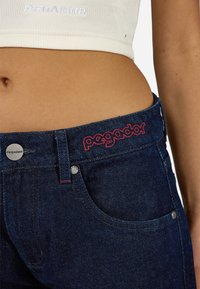 A sötétkék farmernadrágon piros hímezett logó található a derékrészen, ezüst színű fém részekkel és szabványos zsebekkel. A pamut anyag textúrája látható.