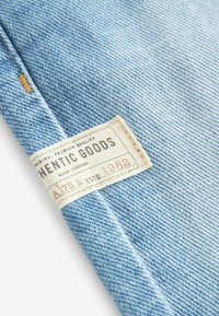 Tissu en denim bleu clair avec un bord replié et une étiquette beige cousue portant l'inscription "AUTHENTIC GOODS" et "Since 1882."