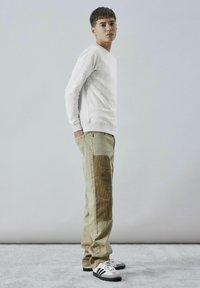 Helles graues Sweatshirt, beige Hose mit strukturiertem braunem Seitenpanel und schwarz-weißen Sneakers. Model steht auf einer hellen Fläche.