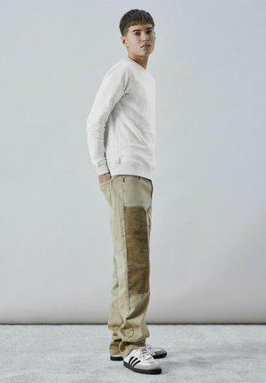 Sweatshirt gris clair, pantalon beige avec une bande latérale texturée marron, et baskets noir et blanc. Modèle se tenant sur une surface claire.