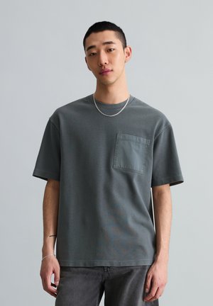 Junger Mann mit kurzen Haaren, der ein dunkelgraues T-Shirt mit Tasche, eine silberne Kette und ein Armband vor einem einfarbigen hellgrauen Hintergrund trägt.