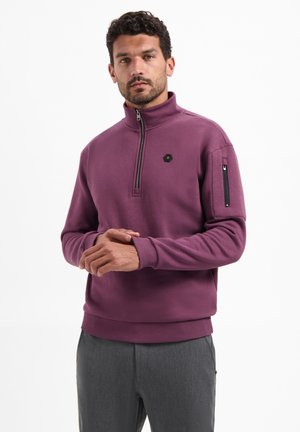 Paarse trui met een hoge kraag, een deels ritssluiting aan de voorkant en een zijzak. Zacht van textuur, met geribbelde manchetten en zoom, en een klein zeshoekig logodetail.