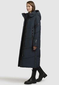 Lang mørkeblå puffjakke med hætte, der har lynlås lukning, sidelommer og knapdetaljer. Jakken har et quiltet design og en lige pasform.