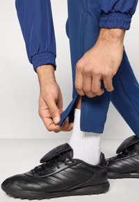 Μπλε αθλητικό παντελόνι με ελαστικά μανσέτα, που διαθέτει πλαϊνή τσέπη, είναι μερικώς ανοιχτό με φερμουάρ. Μαύρα ποδοσφαιρικά παπούτσια με υφή στο επάνω μέρος και λευκές κάλτσες ορατές.