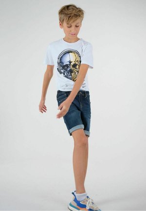 T-shirt blanc avec un design de crâne graphique en or et argent, associé à un short en denim bleu foncé et des chaussures de sport colorées.
