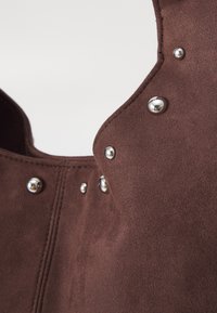 Matériau en daim brun avec des clous-pression argentés le long des bords, possédant une texture lisse et une forme distincte mettant en avant un design structuré.