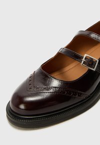 Brown leather Mary Jane shoe with two adjustable straps, round toe, brogue detailing, and a textured rubber sole.