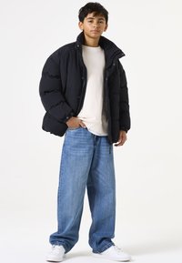 Chaqueta acolchada negra con cuello alto, combinada con unos vaqueros de denim azul holgados. Una camiseta blanca debajo y zapatillas blancas completan el look.