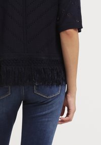 Osoba nosząca granatową bluzkę z krótkim rękawem i frędzlami na dole oraz ciemnoniebieskie dżinsy, widoczna od talii z tyłu do połowy uda.