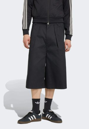 Osoba ubrana w czarne szerokie, krótkie spodnie, czarną kurtkę na zamek z białymi paskami na rękawach, czarne skarpetki Adidas oraz czarne buty sportowe Adidas.