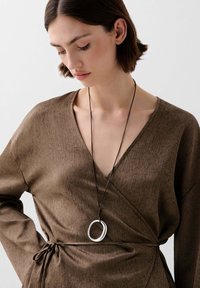 Vestido marrón de tela texturizada, con un profundo escote en V y cintura ajustada con lazo. Un colgante circular de plata cuelga de un collar decordón oscuro.