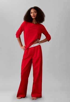 Mujer con cabello rizado viste una camisa de manga larga roja y pantalones rojos sueltos, descalza, posando con una mano en la cadera y la otra levantando ligeramente la camisa.