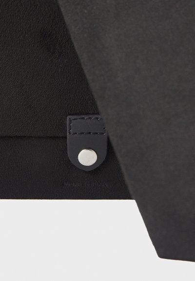 Et produkt i svart semsket skinn med en glatt overflate, en lærstropp med trykknappslukking og preget tekst som indikerer "MADE IN INDIA."