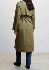 Olijfgroene trenchcoat met een tailleceintuur, plooien aan de achterkant en schouderepauletten, gecombineerd met donkere spijkerbroeken en zwarte schoenen.