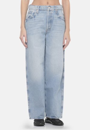 Persona che indossa jeans a vita alta, ampi e di colore azzurro chiaro, e scarpe slip-on nere con frange, in piedi contro uno sfondo bianco.