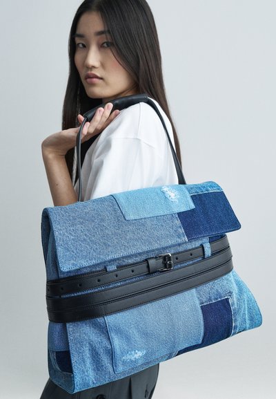 Γυναίκα με μακριά σκούρα μαλλιά κρατά μια μεγάλη τσάντα από πατσαβούρια τζιν με μαύρη δερμάτινη λαβή στον ώμο της, μπροστά σε απλό φόντο.