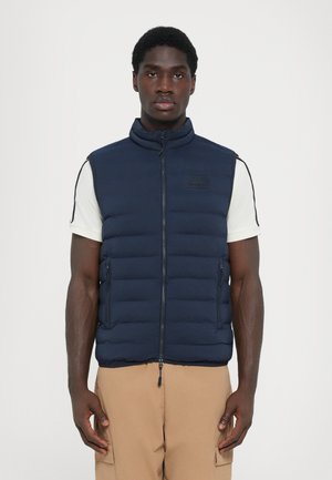 Navy puffer vest with a high collar, quilted design, two front zip pockets, and a label on the chest, worn over a white shirt.