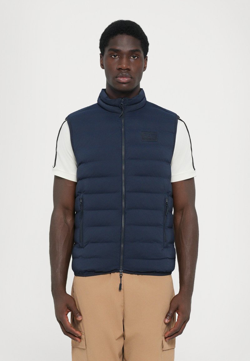 Navy puffer vest with a high collar, quilted design, two front zip pockets, and a label on the chest, worn over a white shirt.