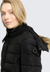 Zwart pufferjack met een hoge kraag, bontgevoerde capuchon, gequiltde textuur en drukknoopdetail op de mouw. Ritsluiting aan de voorkant.