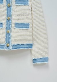 Chaqueta de crema texturizada con detalles de mezclilla azul deshilachados y cuatro bolsillos con parche. Los cierres de botones dorados añaden un detalle decorativo.