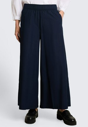 Pantalon large bleu marine avec taille élastique, poches latérales, porté avec des mocassins noirs et un chemisier blanc à manches bouffantes.