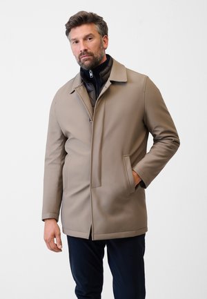 Manteau imperméable beige à coupe droite, avec col montant et poches à rabat. Porté par-dessus une couche intérieure noire à fermeture éclair. Texture lisse.