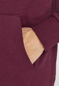 Sudadera con capucha color granate con bolsillo delantero; puños y dobladillo acanalados. El material parece suave con una textura lisa, diseñada para uso casual.