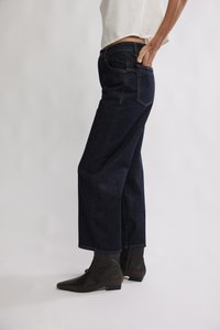 Jean denim bleu foncé à jambes larges, taille haute, avec une conception classique à cinq poches et des détails de couture subtils, associé à des bottines noires.